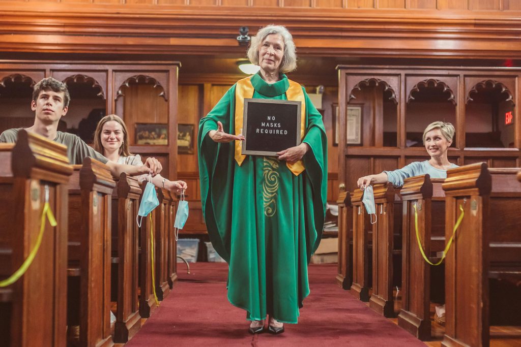 A church service where mask policies have changed, featuring a priest with a sign.